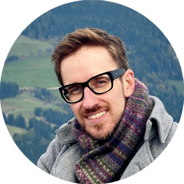A close portrait of me looking straight into the camera. In the background are hills partially covered with forest. I’m wearing a light-gray melton jacket, a colorful scarf, and thick-frame black glasses.